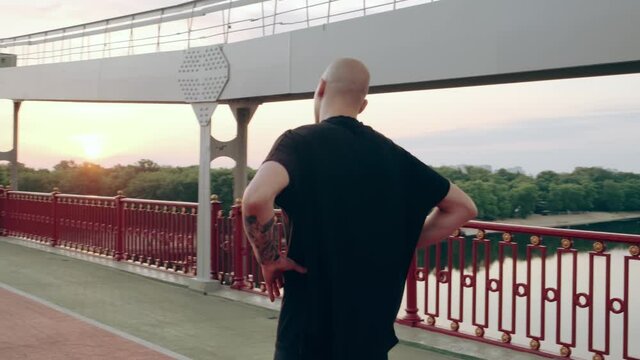 Man runner in black sports uniform doing warm-up before jogging on the pedestrian bridge at sunrise. High quality 4k footage