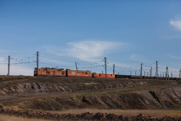 Train with orange locomotive and wagon in steppe. Platform cars for coal and ore transportation. Brown ground. Electric towers, wires, blue sky with cloud. Sunset view.