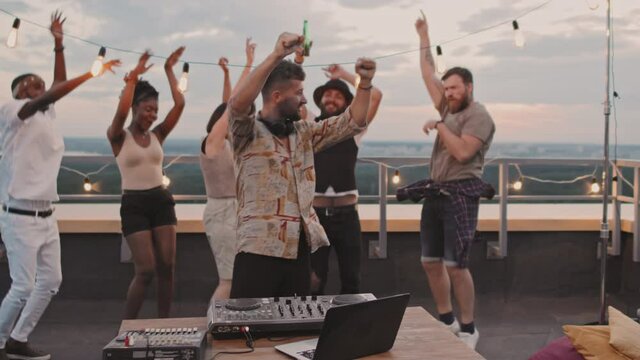 Handheld slowmo shot of male DJ playing set and dancing with group of young people on rooftop terrace on summer evening