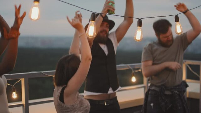 Handheld tracking slowmo shot of group of young friends jumping and dancing at party on rooftop terrace on summer evening