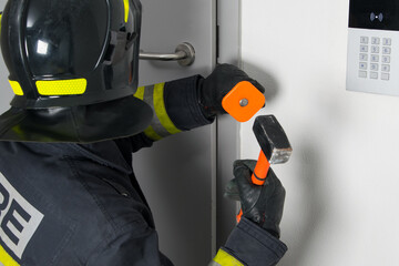 close-up of a firefighter in protective clothing and a hard hat trying to open a door with a...