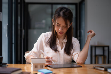 Architect woman using a digital tablet application to hand-draw a design house with a model on a wooden desk. Property idea in real estate.