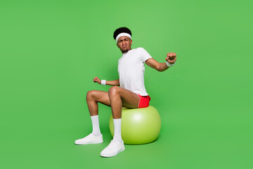 Full length photo of serious funny dark skin guy dressed sport outfit sitting fitball warming up isolated green color background