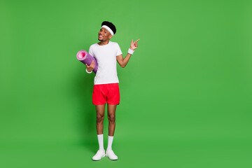 Fototapeta na wymiar Full length photo of charming sweet dark skin man wear white t-shirt holding mat pointing empty space isolated green color background