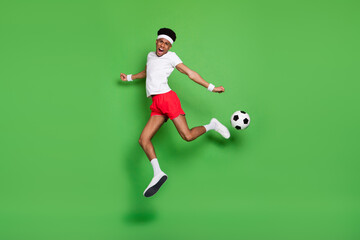 Full length photo of excited pretty dark skin guy dressed sport outfit jumping playing football isolated green color background