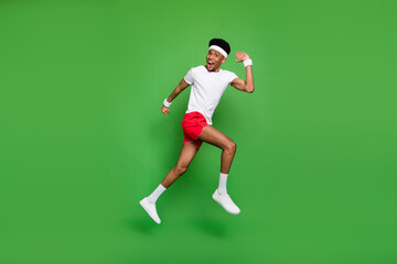 Plakat Full length photo of excited funny dark skin man wear white t-shirt jumping high running fast isolated green color background