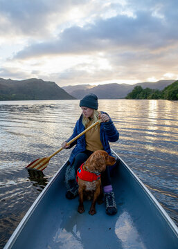 Travel lifestyle view of girl and adventure dog canoeing in red Canadian canoe on Ullswater Lake in Lake District National Park, England, UK.