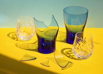 broken transparent empty blue cup among other crystal glasses on yellow background, abstract accident or problem concept, shards of broken glass under hard light
