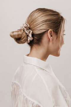 Portrait of stylish blonde with hair collected in silk beige scrunchie, wearing white blouse and posing against light background. Beauty and tenderness concept