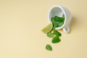 Green leaves of fresh mint and a slice of lime are poured out of a white tea cup on a light plain beige surface