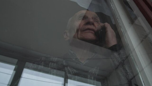 Concentrated Old Man Talks On Smartphone Looking Outside Outdated Window With Bright Sunlight Reflections Low Angle Shot