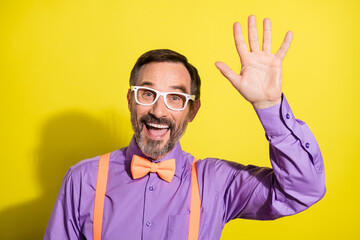 Photo of mature cheerful man happy positive smile waving hand hello greetings isolated over yellow...