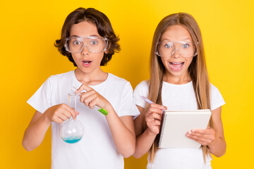 Portrait of two attractive amazed cheerful kids making chemical experiment writing note isolated over bright yellow color background