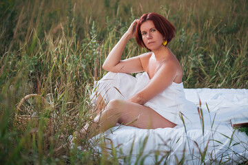 Charming caucasian woman sitting in the white bed in a field at sunset in summer