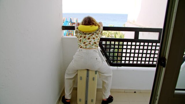 a tourist says goodbye to the sea, rest and vacations. a traveler wants to go to the sea sitting in quarantine. a woman with a neck pillow sits on a suitcase in a funny pose on the hotel balcony. a