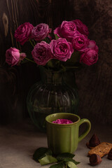 a bouquet of scarlet roses in a transparent vase on a gray table; next to it there is a green ceramic mug, in which a red rose bud is floating. Romantic setting, greeting card