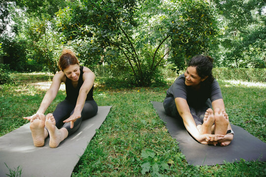 Smiling ethnic female instructor doing Paschimottanasana A yoga pose during training with beginner in park