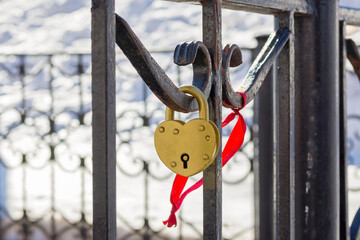 A heart-shaped lock on a wrought-iron fence next to a red ribbon. A symbol of a happy married life. Space for the text. Close up. Horizontal layout.