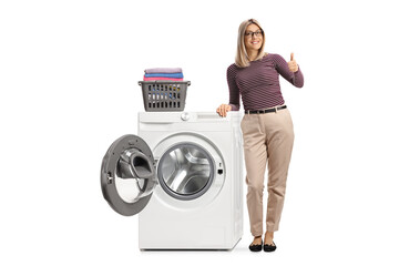 Full length portrait of a blond woman leaning on a washing machine with folded clothes and showing...