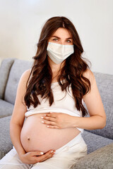 Young beautiful pregnant woman in medical surgical mask and white t-shirt quarantined at home by the window. Coronavirus, virus