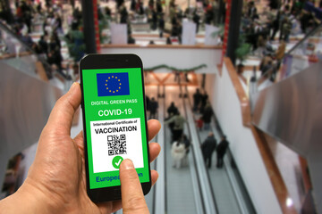 A man in a shopping mall holds a smartphone with the European Union digital green pass for Covid-19 in his hand. Safety concept for Coronavirus and the Green pass - obrazy, fototapety, plakaty