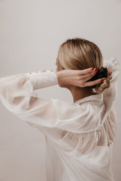 Photo From Back Of Young Tender Girl With Blonde Hair, Black Scrunchie And White Silk Shirt, Adjusting Hairstyle And Posing Isolated Over Light Background