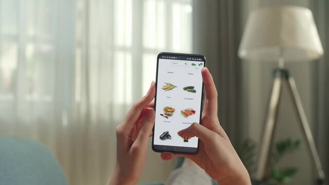 Woman Lying On Couch Using Smartphone Buys In Internet Shop. Female Hand Selects Vegetables In Grocery Online Store
