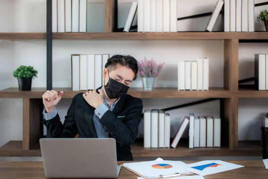 Young asian businessman in suit wearing mask feeling tired and aching have office syndrome and stress while working on laptop computer In the situation of COVID-19 or the coronavirus is spreading.