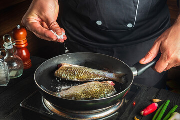 Professional chef prepares fresh fish in pan sprinkling salt with the ingredients. Preparing to cook fish food. Working environment in the restaurant kitchen.