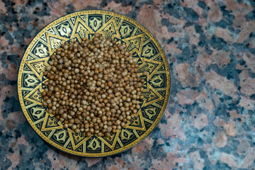 Coriander seeds on metal tray with oriental (middle eastern) motifs, flat lay on a marble board texture.