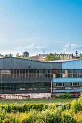 Fototapeta na wymiar Old, metal, rusted factory hall situated in industrial district in a city. Tall grass and bushes in the foreground, cityscape and blue sky in the background.
