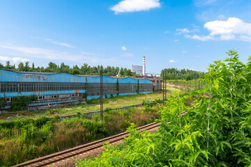 Fototapeta na wymiar Railroad tracks situated between old, rusted factory hall and a slope covered by trees and bushes. Trees and power station with high chimney in the background.