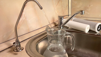 Pouring filtered water into glass pitcher from water filter. Closeup of jug, sink and faucet....