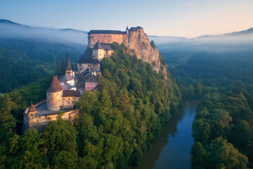 Fototapeta na wymiar Aerial view of Orava Castle situated on a high rock above Orava river, illuminated by rising sun, surrounded by deep forest and foggy mountains. Castles of Slovakia.