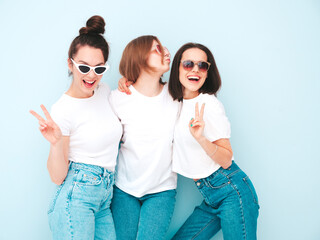 Fototapeta premium Three young beautiful smiling hipster female in trendy same summer white t-shirt and jeans clothes. Sexy carefree women posing near light blue wall in studio. Cheerful and positive models having fun