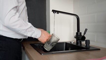 Unrecognizable man washes a mug in the kitchen sink. A jet of water flows from a black tap. The male is dressed in a white shirt and trousers

