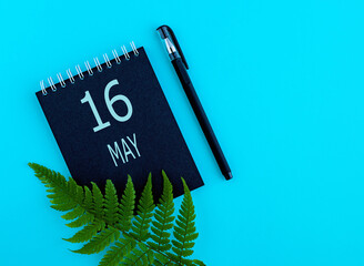 May 16th. Day 16 of month, Calendar date. Black notepad sheet, pen, fern twig, on a blue background. Spring month, day of the year concept