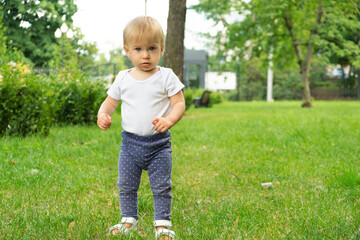 Funny cutest blonde caucasian baby girl of 1 year old, standing on lawn, looking at camera.Infant, spending summertime outdoors in park. Kid and nature concept.Copy space for text