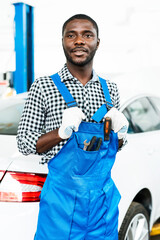 Handsome african american male auto mechanic in overalls and gloves stands in a car showroom against the backdrop of a car