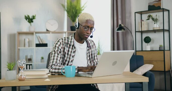 Handsome confident busy young african american man in headphones sitting in front of computer during video conference with client or coworker from home office