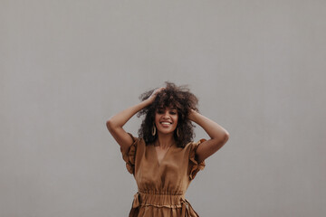 Happy woman in brown dress ruffles hair outside. Curly dark-skinned lady in beige outfit smiles and...