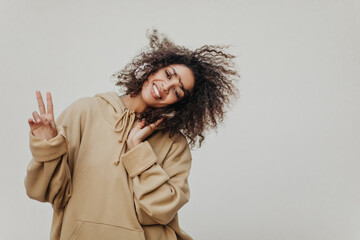 Brown-eyed African girl shows peace sign and dances on grey background. Cheerful woman in oversized beige hoodie listens to music in headphones.