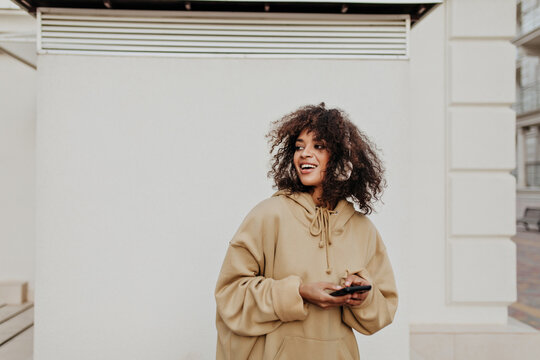 Brunette woman in beige hoodie listens to music in headphones outside. Curly dark-skinned girl holds phone.