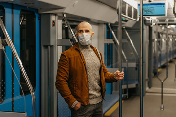 A bald man with a beard in a face mask is holding the handrail in a subway car.