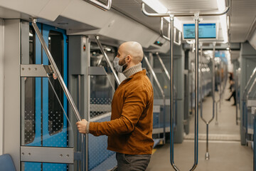 A bald man with a beard in a face mask is holding the handrail in a subway car.