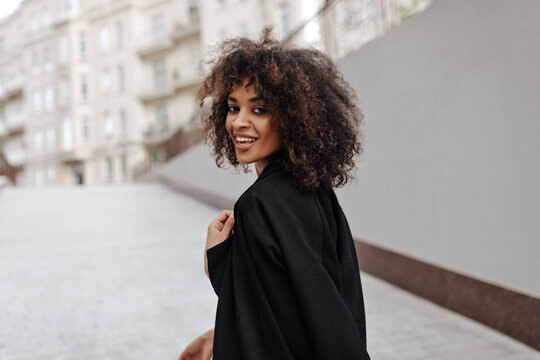 Attractive African woman in black coat walks outside. Dark-skinned curly lady looks into camera and smiles outdoors.