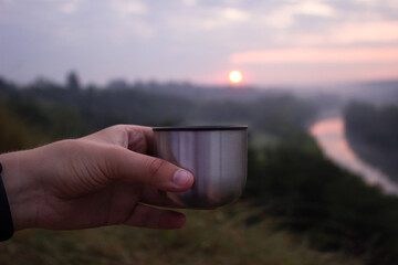 Obraz premium A man holds a metal mug in his hand in the morning against the background of dawn and the river. Travel concept