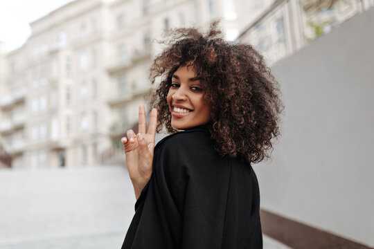 Curly brunette woman in black coat skies peace sign outside. Dark-skinned African lady smiles sincerely and walks outdoors.