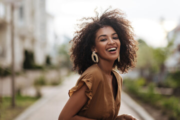 Cheerful brunette woman in brown dress smiles sincerely outdoors. Happy lady laughs and walks in...