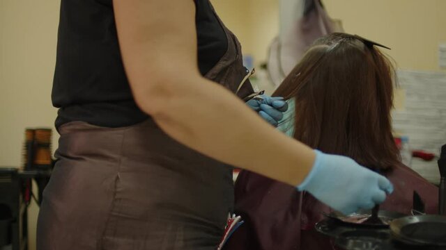 Unrecognizable person dyes hair of young woman in salon. Unknown hairdresser in gloves coloring hair of client in beauty salon. Concept of care and beauty